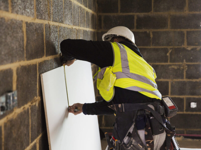 A builder in a hard hat wearing a tool belt, measuring a piece of wood.,Construction Site,England A builder in a hard hat wearing a tool belt, measuring a piece of wood.